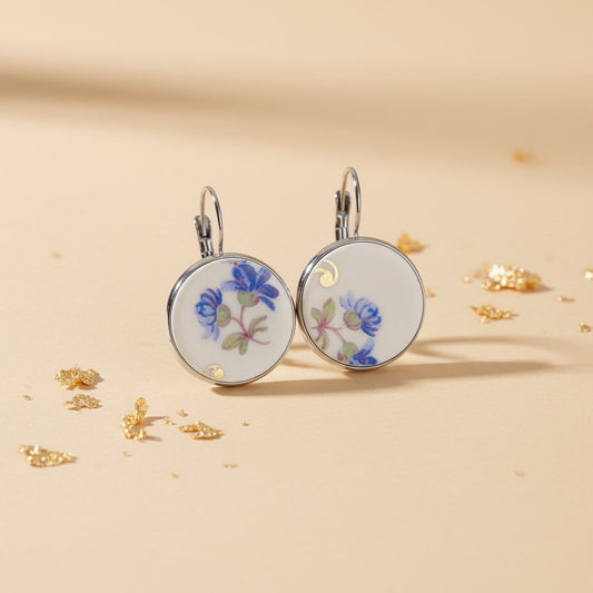 Handgefertigte Porzellanohrringe Modell Caerula von EDELATELIER. Zwei runde Ohrhänger aus weißem Vintage-Porzellan mit einem filigranen, himmelblauen Blüten-Dekor. Die Porzellanelemente sind in silberfarbene Fassungen aus hochwertigem Edelstahl gefasst und hängen an schlichten Ohrhaken. Das Design kombiniert nostalgische Porzellanmalerei mit modernem, minimalistischem Upcycling-Stil.

Bild 2: Detail- und Materiala