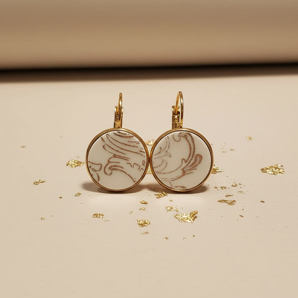Handgefertigte Porzellanohrringe Ornamenta von EDELATELIER. aus upcyceltem antikem Geschirr mit filigranem grauem Ornament-Muster und goldener Fassung auf beigem Hintergrund mit Goldflocken.
