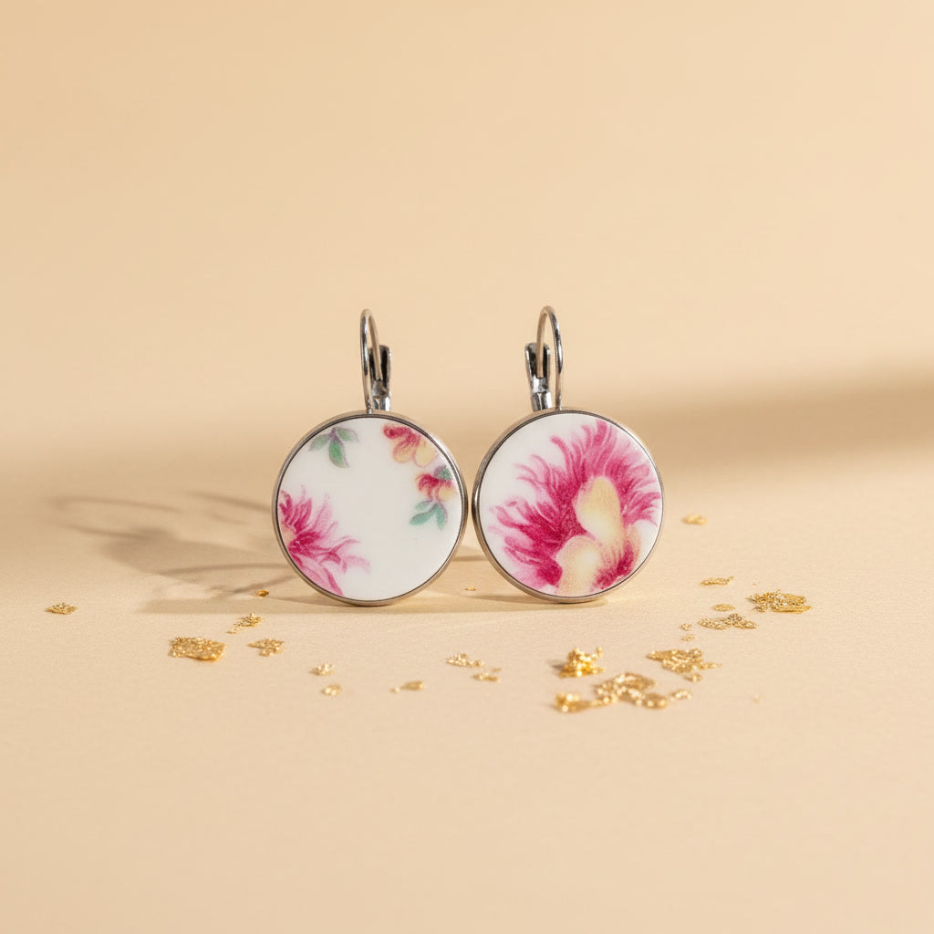 Detailreiche Frontalansicht der runden Porzellanohrringe Petala von EDELATELIER. Das weiße Upcycling-Porzellan zeigt ein lebendiges, historisches Blumenmotiv mit kräftigen roten Blütenblättern und zarten roséfarbenen Schattierungen. Die Fragmente stammen von antikem Geschirr und sind in einer glänzenden, silberfarbenen Fassung eingefasst. Das Schmuckstück liegt auf einem hellbeigen Untergrund, der mit feinen Goldflocken dekoriert ist, was den edlen Manufaktur-Charakter unterstreicht.