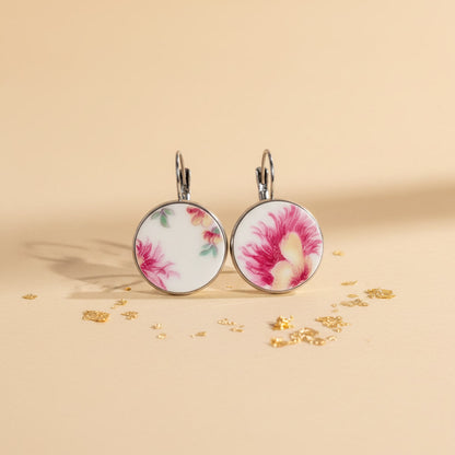 Detailreiche Frontalansicht der runden Porzellanohrringe Petala von EDELATELIER. Das weiße Upcycling-Porzellan zeigt ein lebendiges, historisches Blumenmotiv mit kräftigen roten Blütenblättern und zarten roséfarbenen Schattierungen. Die Fragmente stammen von antikem Geschirr und sind in einer glänzenden, silberfarbenen Fassung eingefasst. Das Schmuckstück liegt auf einem hellbeigen Untergrund, der mit feinen Goldflocken dekoriert ist, was den edlen Manufaktur-Charakter unterstreicht.