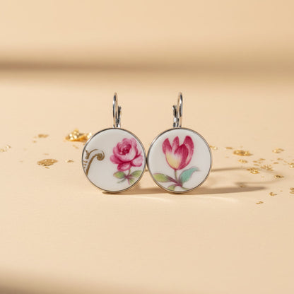 Detailreiche Frontalansicht der Porzellan-Ohrhänger Venusta von EDELATELIER. Das Paar zeigt zwei unterschiedliche, aber harmonierende florale Motive aus antikem Porzellan: eine prachtvolle rote Rose und eine elegante Tulpe in warmen Rottönen mit zarten grünen Blättern. Die runden Upcycling-Elemente sind in glänzende silberfarbene Fassungen gebettet und liegen auf einem hellbeigen Untergrund mit feinen Goldflocken, was das exklusive Design unterstreicht.