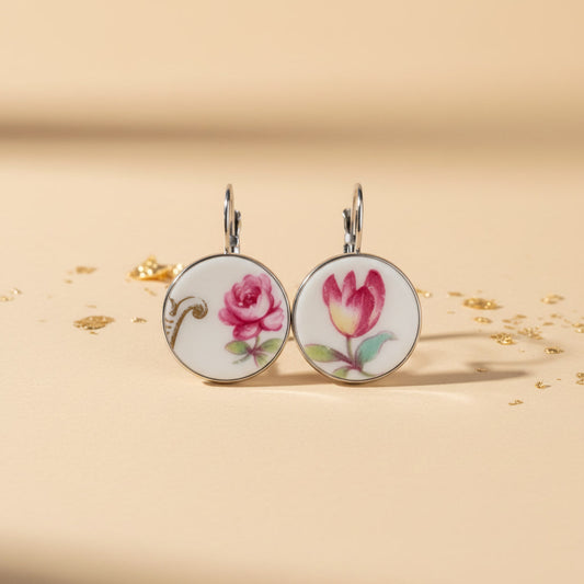 Detailreiche Frontalansicht der Porzellan-Ohrhänger Venusta von EDELATELIER. Das Paar zeigt zwei unterschiedliche, aber harmonierende florale Motive aus antikem Porzellan: eine prachtvolle rote Rose und eine elegante Tulpe in warmen Rottönen mit zarten grünen Blättern. Die runden Upcycling-Elemente sind in glänzende silberfarbene Fassungen gebettet und liegen auf einem hellbeigen Untergrund mit feinen Goldflocken, was das exklusive Design unterstreicht.