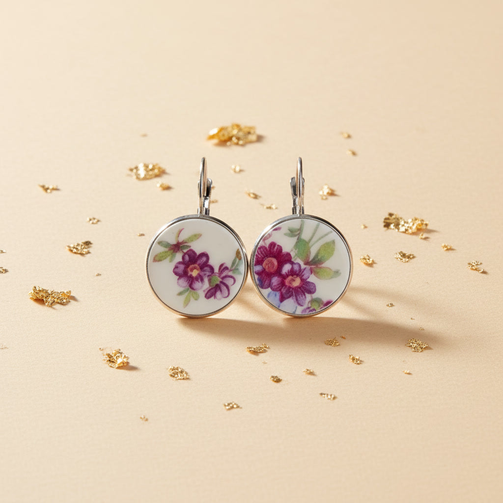 Hochauflösende Frontalansicht der runden Porzellan-Ohrhänger Viola von EDELATELIER. Das Design zeigt ein prachtvolles, violettes Blütenmotiv auf reinweißem historischem Porzellan, das aus antikem Geschirr gewonnen wurde. Die tiefen Purpurtöne der Blumen harmonieren perfekt mit der kühlen, silberfarbenen Fassung. Das Paar liegt auf einem hellen, strukturierten Untergrund mit dezenten Goldflocken-Akzenten.