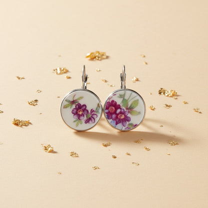 Hochauflösende Frontalansicht der runden Porzellan-Ohrhänger Viola von EDELATELIER. Das Design zeigt ein prachtvolles, violettes Blütenmotiv auf reinweißem historischem Porzellan, das aus antikem Geschirr gewonnen wurde. Die tiefen Purpurtöne der Blumen harmonieren perfekt mit der kühlen, silberfarbenen Fassung. Das Paar liegt auf einem hellen, strukturierten Untergrund mit dezenten Goldflocken-Akzenten.