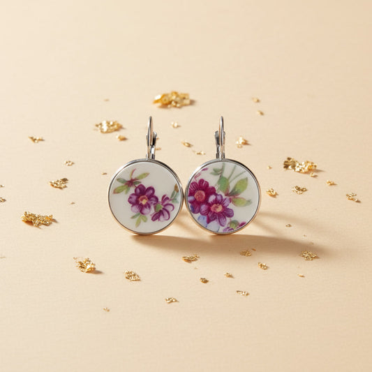 Hochauflösende Frontalansicht der runden Porzellan-Ohrhänger Viola von EDELATELIER. Das Design zeigt ein prachtvolles, violettes Blütenmotiv auf reinweißem historischem Porzellan, das aus antikem Geschirr gewonnen wurde. Die tiefen Purpurtöne der Blumen harmonieren perfekt mit der kühlen, silberfarbenen Fassung. Das Paar liegt auf einem hellen, strukturierten Untergrund mit dezenten Goldflocken-Akzenten.
