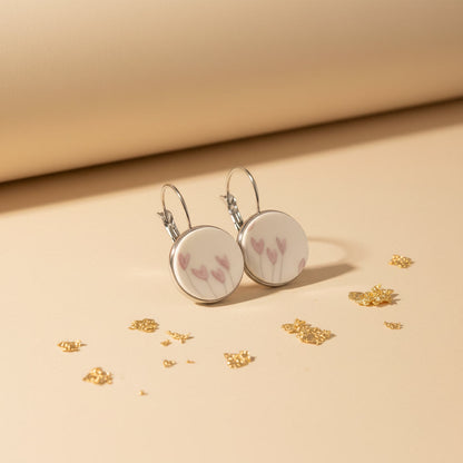 Handgefertigte Porzellanohrringe „Flosculus“ von EDELATELIER mit zartrosa Tulpenmotiven auf weißem Porzellan, eingefasst in silberfarbene Rundfassungen, präsentiert auf beigem Hintergrund mit Goldakzenten.