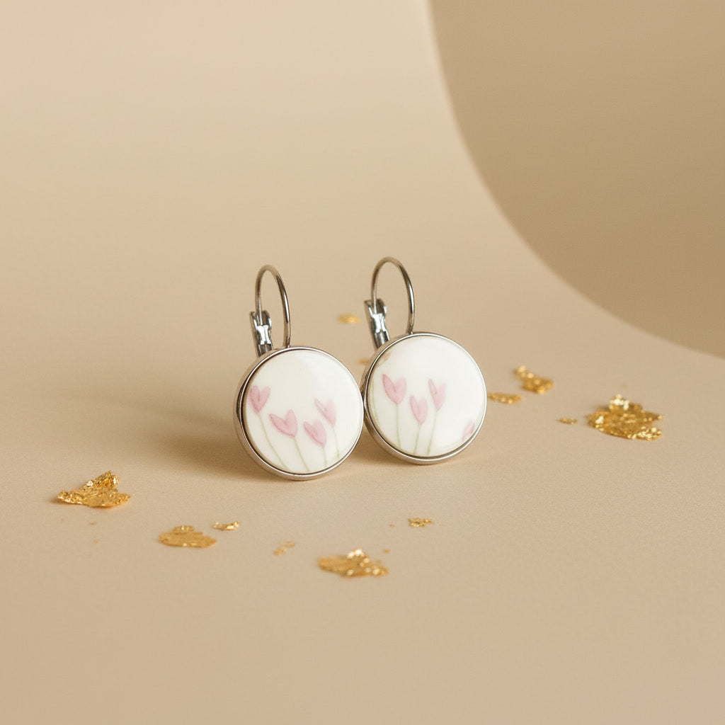 Handgefertigte Porzellanohrringe „Flosculus“ von EDELATELIER mit zartrosa Tulpenmotiven auf weißem Porzellan, eingefasst in silberfarbene Rundfassungen, präsentiert auf beigem Hintergrund mit Goldakzenten.