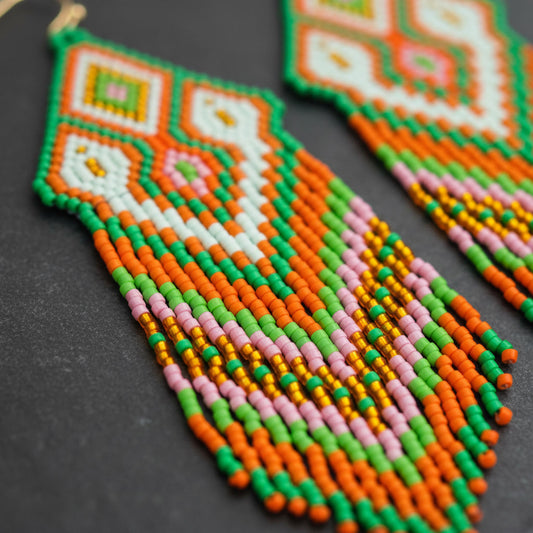 Detailaufnahme der handgewebten Statement-Ohrringe Primavera Viva von EDELATELIER. aus einer schrägen Perspektive auf dunklem Naturstein, die das präzise geometrische Miyuki-Perlenmuster in Grün, Orange und Pink hervorhebt.