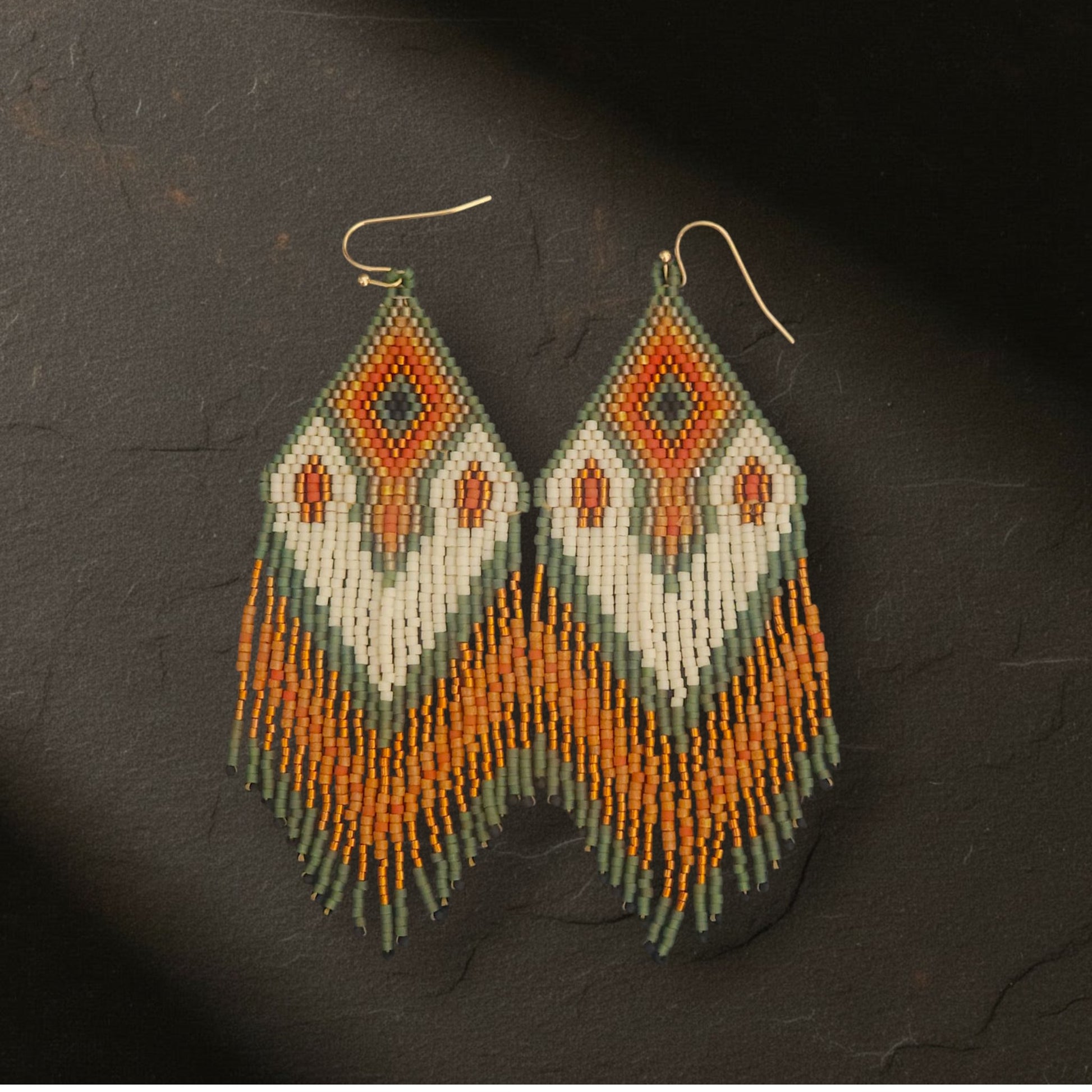 Ein Paar handgefertigte Statement-Ohrringe Modell Raggio di Fiammetta von EDELATELIER, präsentiert auf dunklem Naturschiefer. Die 10 cm langen Ohrringe zeigen ein leuchtendes Farbspiel aus Orange, Salbeigrün und Creme mit goldenen Akzenten. Die filigranen Perlenfransen und das präzise Webmuster kommen auf dem rustikalen Steinuntergrund kontrastreich zur Geltung.