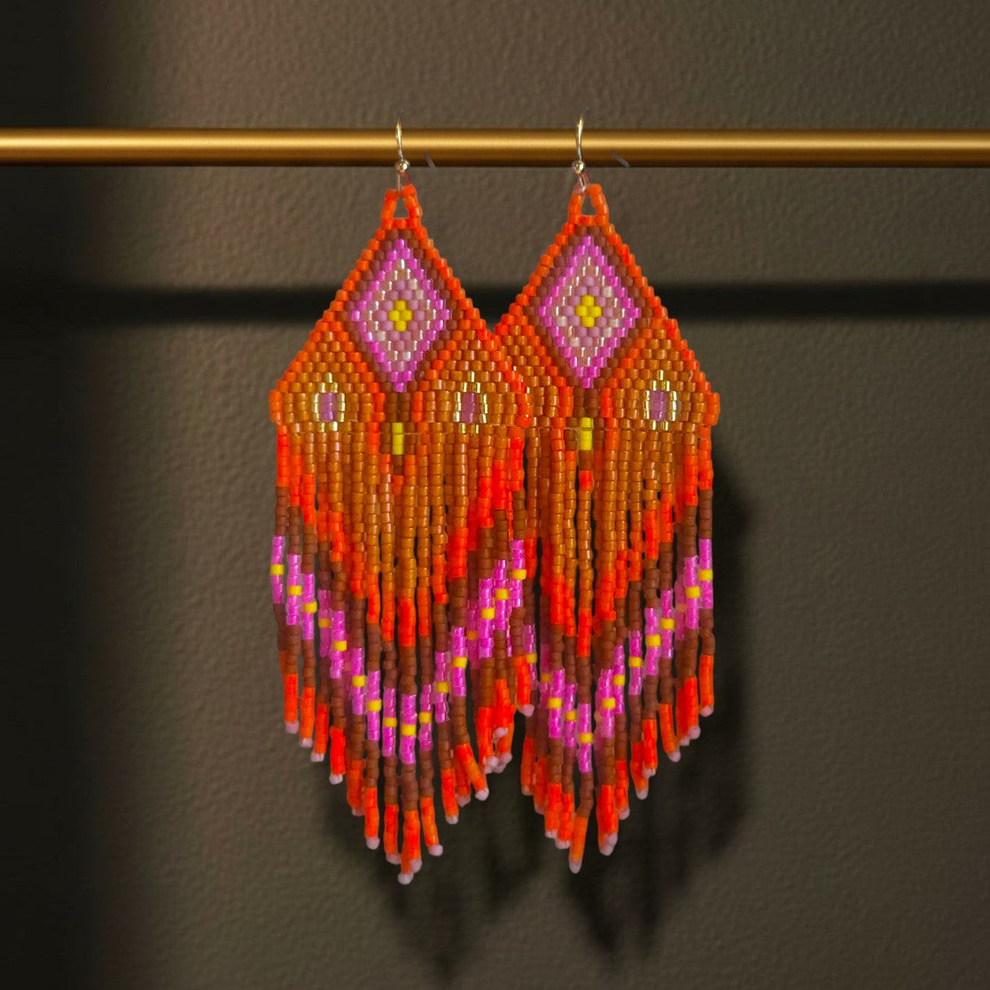 Ein Paar handgefertigte Statement-Ohrringe Modell Riva Ardente von EDELATELIER, präsentiert an einer goldenen Stange. Die 10 cm langen Ohrringe bestechen durch ein lebendiges Muster in kräftigem Orange, leuchtendem Pink und warmem Braun mit gelben Akzenten.