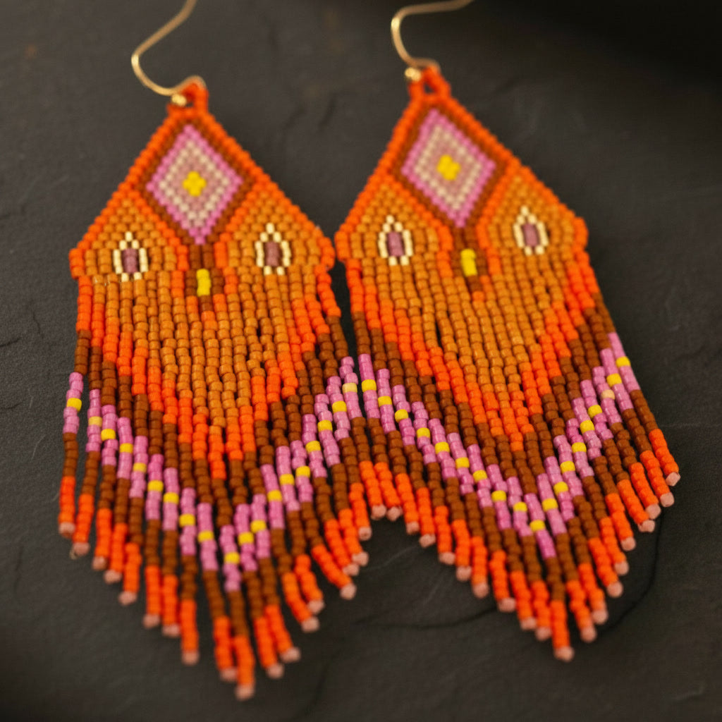 Eine detaillierte Makroaufnahme der "Riva Ardente" Ohrringe auf einer dunklen Schieferplatte. Die präzise gewebten Miyuki-Perlen in kräftigem Orange und Magenta-Pink zeigen ein kunstvolles Webmuster mit feinen Farbübergängen.