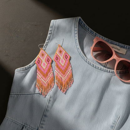 Ein stimmungsvolles Produktfoto im Flatlay-Stil zeigt die handgefertigten Perlenohrringe "Rosa Antico" von EDELATELIER. auf einem hellblauen Denim-Kleid. Die langen Fransenohrringe in Rosa-, Zimt- und Cremetönen liegen auf der Schulterpartie des Kleides. Daneben ist eine rosa Sonnenbrille platziert, die den sommerlichen Look unterstreicht. Ein diagonaler Schattenwurf sorgt für eine hochwertige Lichtstimmung und hebt die feine Struktur der gewebten Perlen hervor.