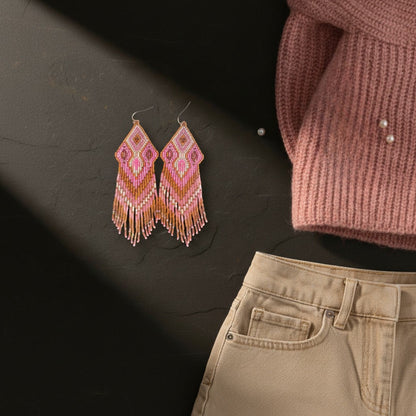 Auf einer dunklen Schieferplatte liegen die rosa-zimtfarbenen Ohrringe. Rechts ragt ein altrosa Strickpullover mit kleinen Perlenapplikationen ins Bild, während unten der Bund einer beigen Jeans zu sehen ist.