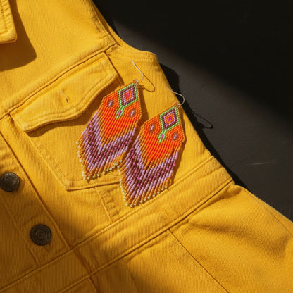 Die pink-orangefarbenen Perlenohrringe sind dekorativ auf einem senfgelben Jeanskleid platziert. Der Kontrast zwischen den warmen Farben des Stoffes und den leuchtenden Schmuckfarben unterstreicht den modernen Boho-Stil.