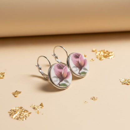 Detailaufnahme der handgefertigten Porzellanohrringe Rosa Splendida von EDELATELIER aus einer schrägen Seitenperspektive. Die beiden runden Ohrhänger stehen aufrecht auf einem beigefarbenen Untergrund, der mit dekorativen Goldflocken bestreut ist. Diese Ansicht betont die flache, elegante Form der Porzellanscherben und die hochwertige Einfassung in silberfarbene Fassungen. Die silbernen Ohrhaken (Brisuren) sind deutlich in ihrer geschwungenen Form erkennbar.