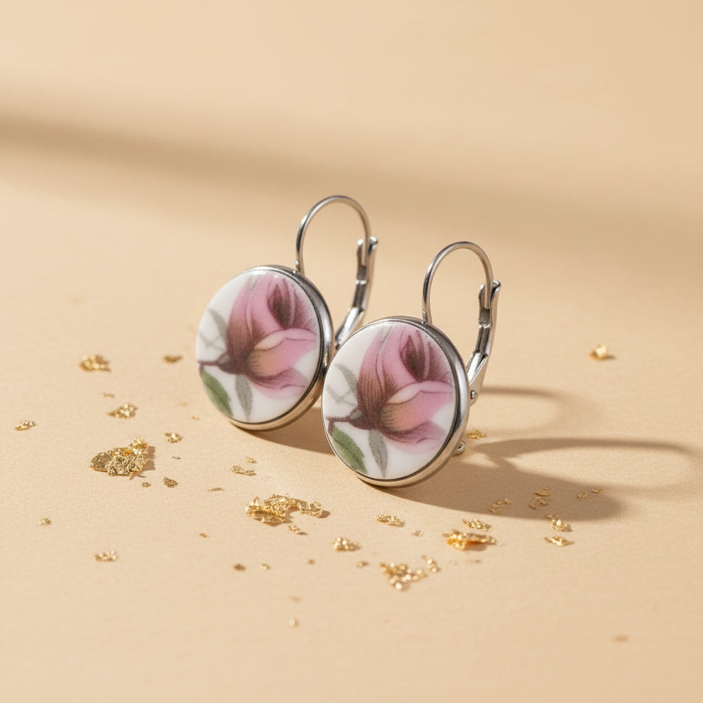 Detailaufnahme der handgefertigten Porzellanohrringe Rosa Splendida von EDELATELIER aus einer schrägen Seitenperspektive. Die beiden runden Ohrhänger stehen aufrecht auf einem beigefarbenen Untergrund, der mit dekorativen Goldflocken bestreut ist. Diese Ansicht betont die flache, elegante Form der Porzellanscherben und die hochwertige Einfassung in silberfarbene Fassungen. Die silbernen Ohrhaken (Brisuren) sind deutlich in ihrer geschwungenen Form erkennbar.