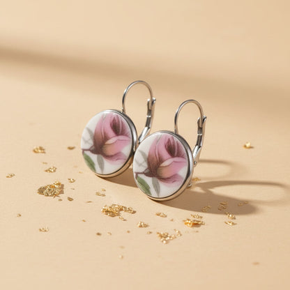 Detailaufnahme der handgefertigten Porzellanohrringe Rosa Splendida von EDELATELIER aus einer schrägen Seitenperspektive. Die beiden runden Ohrhänger stehen aufrecht auf einem beigefarbenen Untergrund, der mit dekorativen Goldflocken bestreut ist. Diese Ansicht betont die flache, elegante Form der Porzellanscherben und die hochwertige Einfassung in silberfarbene Fassungen. Die silbernen Ohrhaken (Brisuren) sind deutlich in ihrer geschwungenen Form erkennbar.