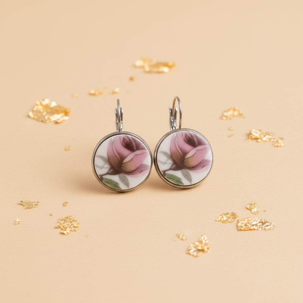 Ein Paar handgefertigte Porzellanohrringe von EDELATELIER, präsentiert vor einem hellen, neutralen Hintergrund. Die runden Ohrhänger zeigen jeweils eine detaillierte, rosafarbene Rosenknospe in klassischer Porzellanmalerei. Die Blütenblätter sind in verschiedenen Nuancen von Altrosa und kräftigem Pink schattiert, ergänzt durch feine grüne Blätter und bräunliche Stiele. Das reinweiße Porzellan ist in eine silberfarbene Fassung eingefasst, die bündig mit der Oberfläche abschließt. 