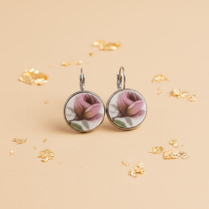 Ein Paar handgefertigte Porzellanohrringe von EDELATELIER, präsentiert vor einem hellen, neutralen Hintergrund. Die runden Ohrhänger zeigen jeweils eine detaillierte, rosafarbene Rosenknospe in klassischer Porzellanmalerei. Die Blütenblätter sind in verschiedenen Nuancen von Altrosa und kräftigem Pink schattiert, ergänzt durch feine grüne Blätter und bräunliche Stiele. Das reinweiße Porzellan ist in eine silberfarbene Fassung eingefasst, die bündig mit der Oberfläche abschließt. 
