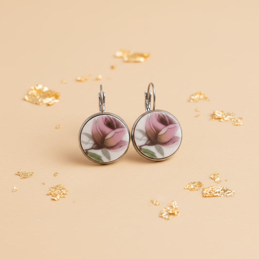 Ein Paar handgefertigte Porzellanohrringe von EDELATELIER, präsentiert vor einem hellen, neutralen Hintergrund. Die runden Ohrhänger zeigen jeweils eine detaillierte, rosafarbene Rosenknospe in klassischer Porzellanmalerei. Die Blütenblätter sind in verschiedenen Nuancen von Altrosa und kräftigem Pink schattiert, ergänzt durch feine grüne Blätter und bräunliche Stiele. Das reinweiße Porzellan ist in eine silberfarbene Fassung eingefasst, die bündig mit der Oberfläche abschließt. 