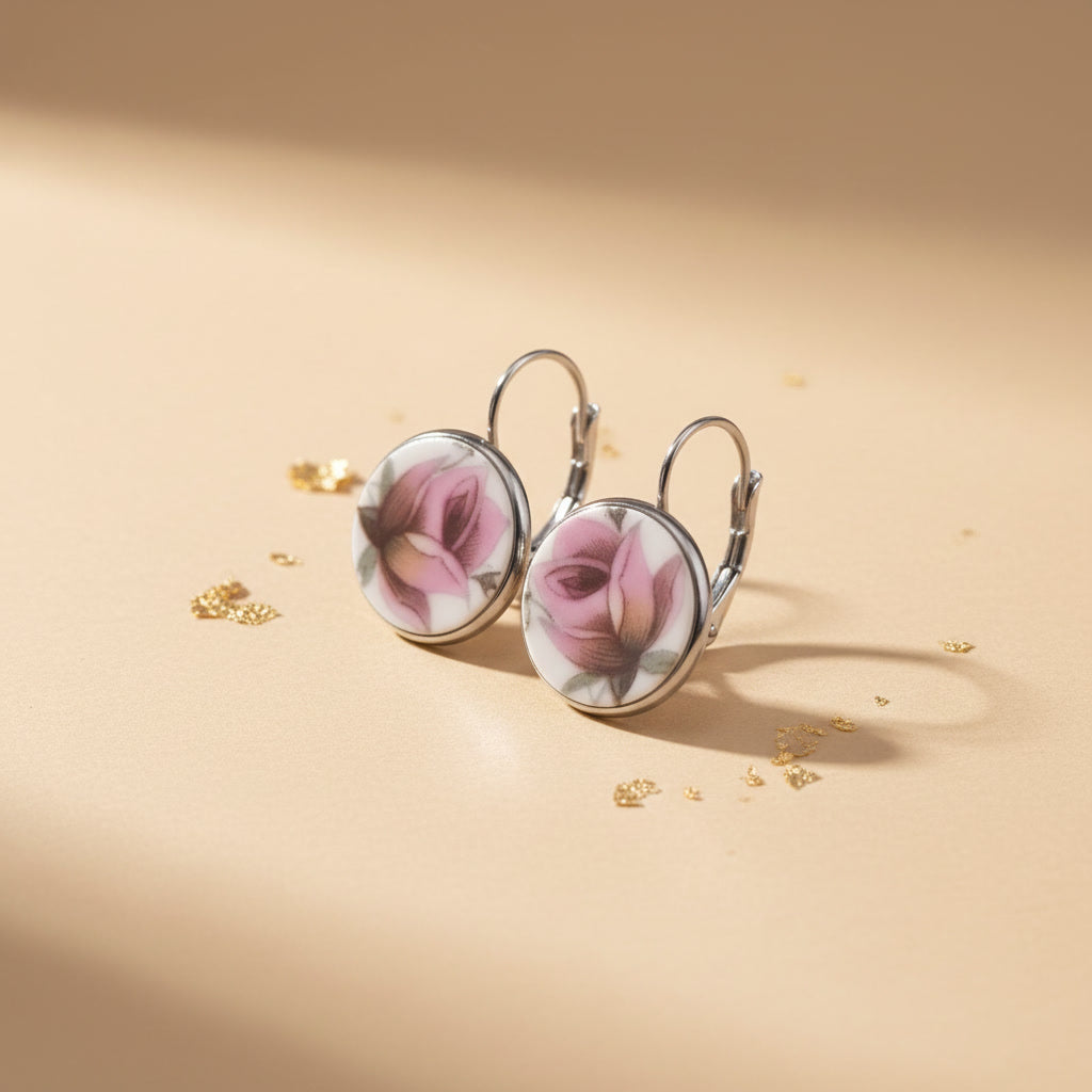 Detailaufnahme der handgefertigten Porzellanohrringe Rosa Splendida von EDELATELIER aus einer schrägen Seitenperspektive. Die beiden runden Ohrhänger stehen aufrecht auf einem beigefarbenen Untergrund, der mit dekorativen Goldflocken bestreut ist. Diese Ansicht betont die flache, elegante Form der Porzellanscherben und die hochwertige Einfassung in silberfarbene Fassungen. Die silbernen Ohrhaken (Brisuren) sind deutlich in ihrer geschwungenen Form erkennbar. Das Licht fällt von rechts ein und erzeugt einen 