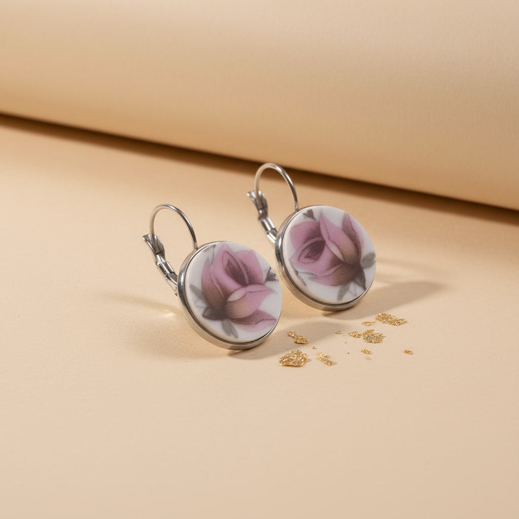 Detailaufnahme der handgefertigten Porzellanohrringe Rosa Splendida von EDELATELIER aus einer schrägen Seitenperspektive. Die beiden runden Ohrhänger stehen aufrecht auf einem beigefarbenen Untergrund, der mit dekorativen Goldflocken bestreut ist. Diese Ansicht betont die flache, elegante Form der Porzellanscherben und die hochwertige Einfassung in silberfarbene Fassungen. Die silbernen Ohrhaken (Brisuren) sind deutlich in ihrer geschwungenen Form erkennbar. Das Licht fällt von rechts ein und erzeugt einen 