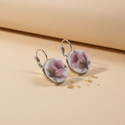 Detailaufnahme der handgefertigten Porzellanohrringe Rosa Splendida von EDELATELIER aus einer schrägen Seitenperspektive. Die beiden runden Ohrhänger stehen aufrecht auf einem beigefarbenen Untergrund, der mit dekorativen Goldflocken bestreut ist. Diese Ansicht betont die flache, elegante Form der Porzellanscherben und die hochwertige Einfassung in silberfarbene Fassungen. Die silbernen Ohrhaken (Brisuren) sind deutlich in ihrer geschwungenen Form erkennbar. Das Licht fällt von rechts ein und erzeugt einen 