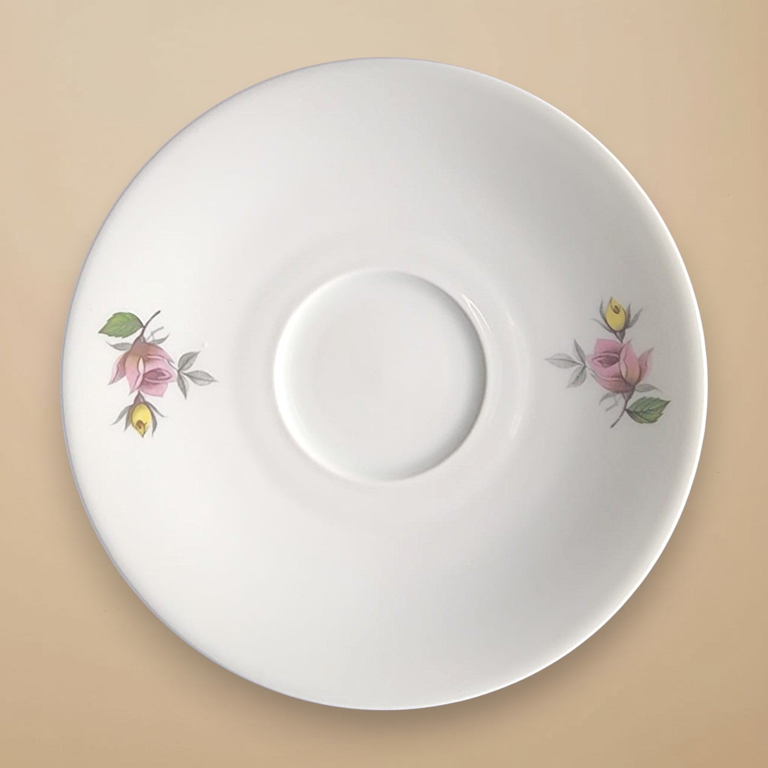Produktfoto des antiken Porzellantellers, der als Ursprung für die Rosa Splendida Ohrringe von EDELATELIER dient. Die Draufsicht zeigt eine reinweiße Untertasse mit einem klassischen floralen Dekor. Links und rechts auf dem Tellerrand ist jeweils eine kunstvoll gemalte, rosa Rosenknospe mit gelben Akzenten und grünen Blättern zu sehen. Das Design ist symmetrisch angeordnet. Der Teller liegt auf einem warmen, beigefarbenen Hintergrund. 