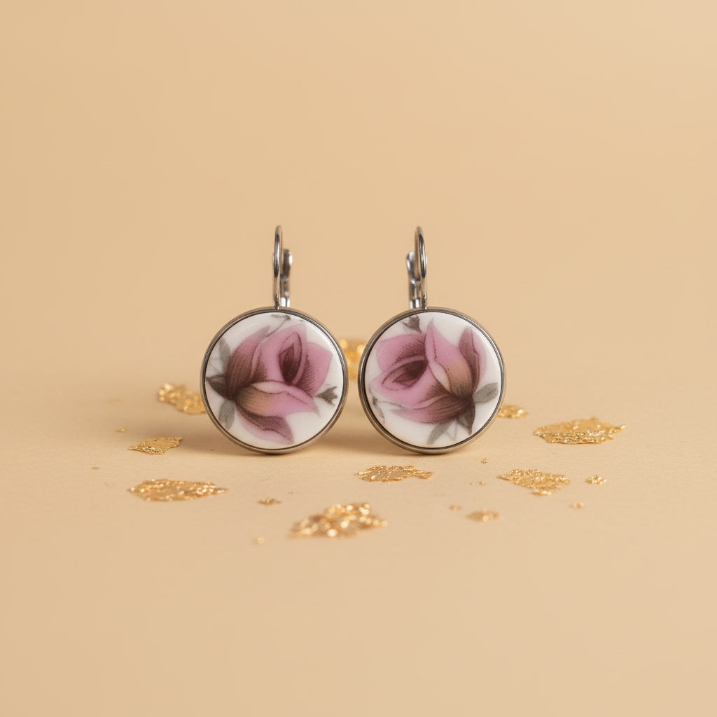 Ein Paar handgefertigte Porzellanohrringe von EDELATELIER, präsentiert vor einem hellen, neutralen Hintergrund. Die runden Ohrhänger zeigen jeweils eine detaillierte, rosafarbene Rosenknospe in klassischer Porzellanmalerei. Die Blütenblätter sind in verschiedenen Nuancen von Altrosa und kräftigem Pink schattiert, ergänzt durch feine grüne Blätter und bräunliche Stiele. Das reinweiße Porzellan ist in eine silberfarbene Fassung eingefasst, die bündig mit der Oberfläche abschließt. Die silbernen Ohrhaken (Bris