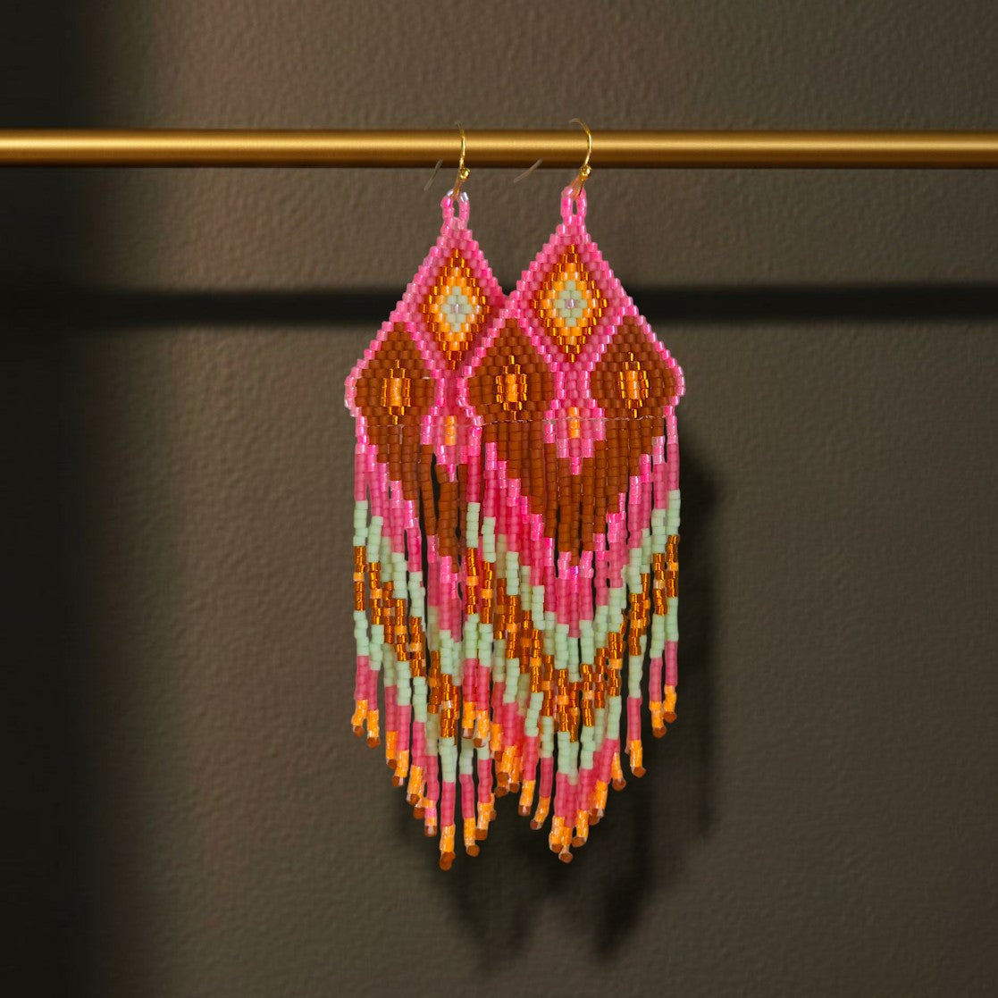 Handgewebte Perlenohrringe Tropico Rosa mit geometrischem Muster in Rosa, Mint und Terrakotta vor dunklem Hintergrund, hängend an einer goldenen Stange. 