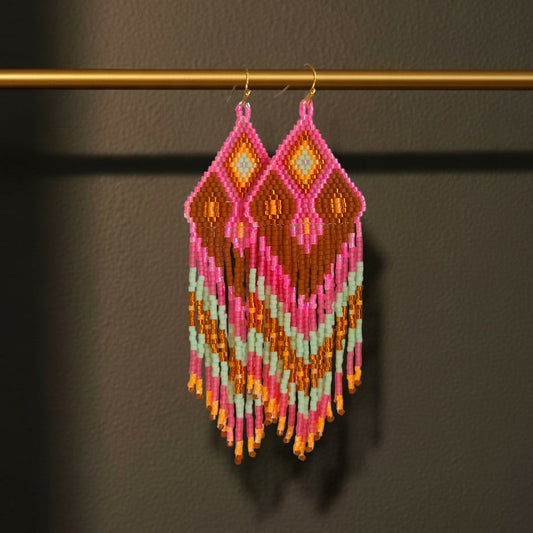 Handgewebte Perlenohrringe Tropico Rosa mit geometrischem Muster in Rosa, Mint und Terrakotta vor dunklem Hintergrund, hängend an einer goldenen Stange. 