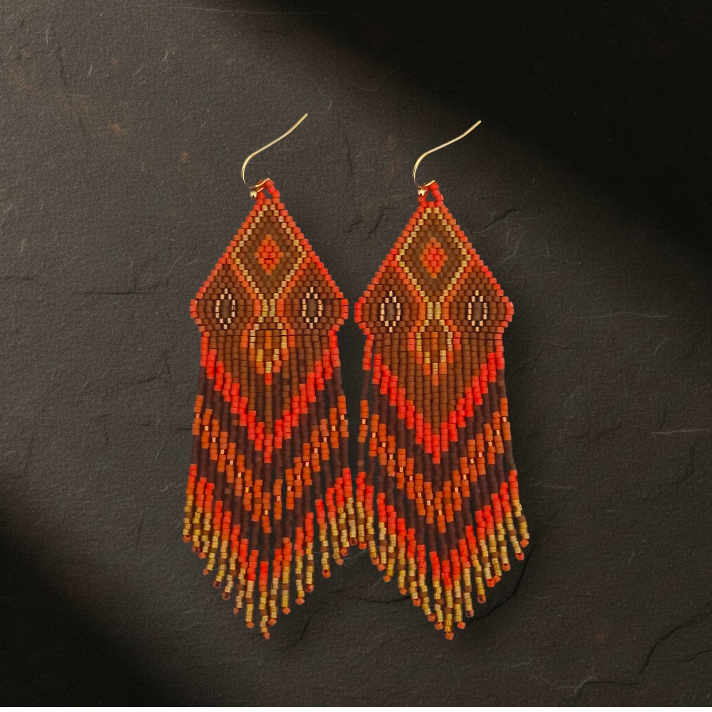 Ein Paar handgefertigte, geometrische Perlentropfen-Ohrringe, die auf einem einem dunklen Untergrund liegen. Der obere Teil ist ein rautenförmiges Muster in leuchtendem Orangerot mit einem kleinen, bronzebraunen Perlen im Inneren, der von goldenen Perlen umrandet ist. Darunter liegen feine Perlenfransen in Schichten von Orangerot, Rostrot, Bronzebraun und Goldgelb, die in einem abgestuften Fransenmuster enden. Der Hintergrund ist ein dunkler Schieferstein.