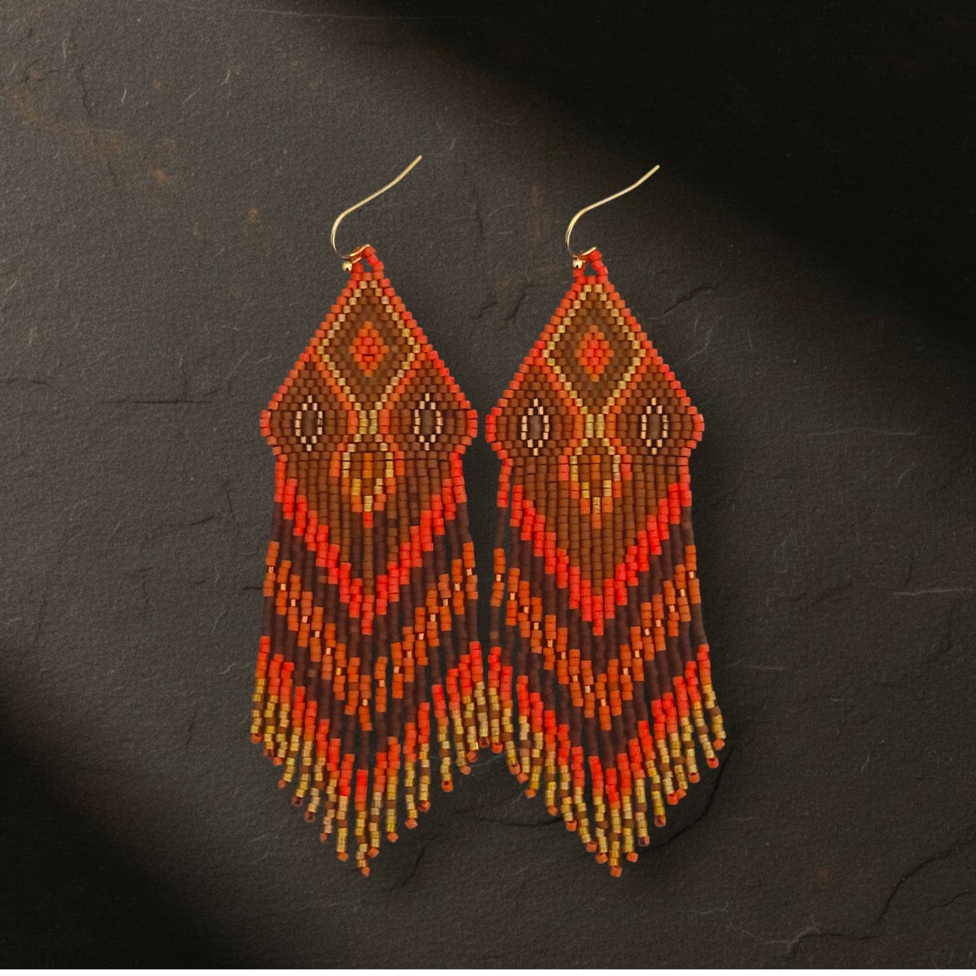 Ein Paar handgefertigte, geometrische Perlentropfen-Ohrringe, die auf einem einem dunklen Untergrund liegen. Der obere Teil ist ein rautenförmiges Muster in leuchtendem Orangerot mit einem kleinen, bronzebraunen Perlen im Inneren, der von goldenen Perlen umrandet ist. Darunter liegen feine Perlenfransen in Schichten von Orangerot, Rostrot, Bronzebraun und Goldgelb, die in einem abgestuften Fransenmuster enden. Der Hintergrund ist ein dunkler Schieferstein.