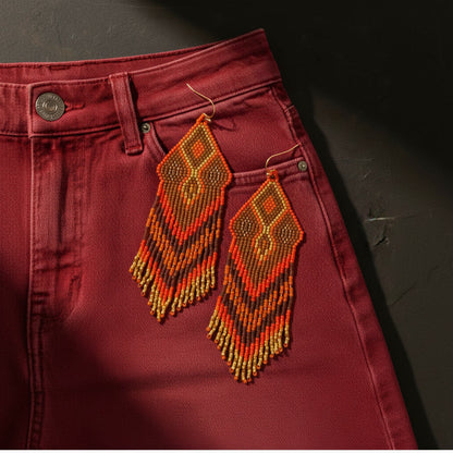 Die rot-orangefarbenen Perlenohrringe liegen dekorativ auf der Tasche einer weinroten Jeans. Der Ton-in-Ton-Effekt mit dem dunklen Stoff unterstreicht die archaische Kraft des Designs.