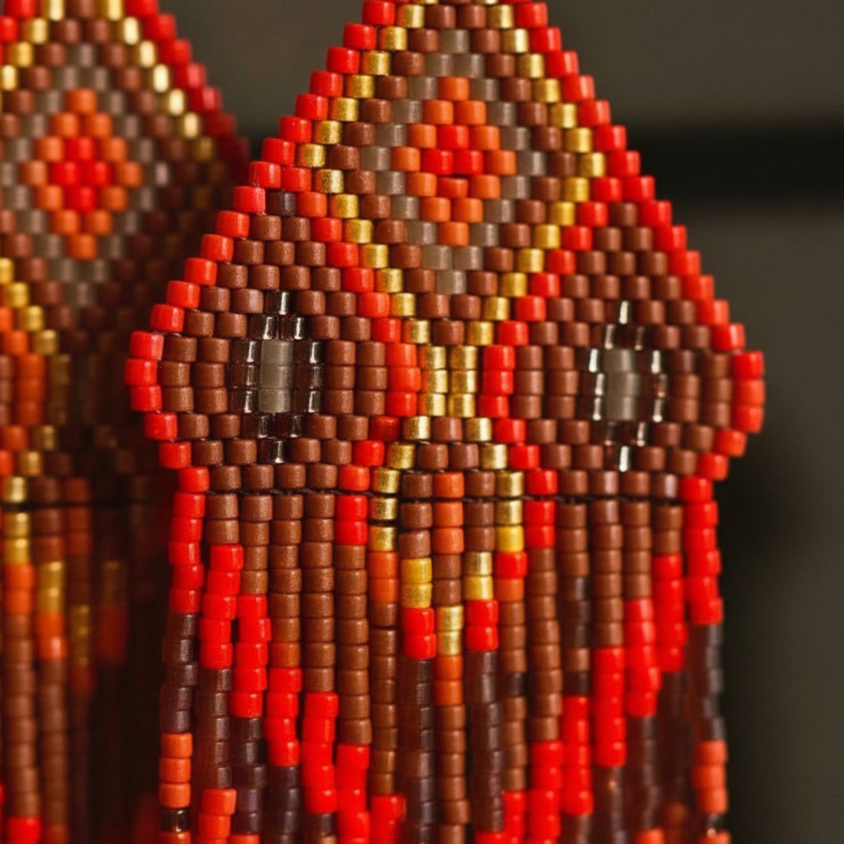Eine detaillierte Makroaufnahme handgewebter Perlenohrringe von EDELATELIER. Das filigrane Muster aus hochwertigen Miyuki-Delica-Perlen zeigt ein geometrisches Rauten-Design in kräftigem Orangerot und warmem Erdbraun, akzentuiert durch glänzende goldene Perlen in der Mitte. Die präzise Anordnung der winzigen Perlen und der Übergang in die feinen, fließenden Fransen am unteren Rand sind deutlich erkennbar.
