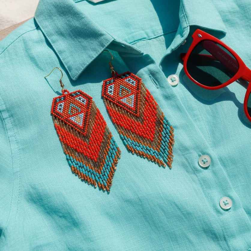 Handgewebte Statement-Ohrringe Rosso di Mare von EDELATELIER. in Rot, Braun und Türkis, arrangiert auf einem türkisfarbenen Leinenhemd neben einer roten Sonnenbrille in einer sommerlichen Produktaufnahme.
