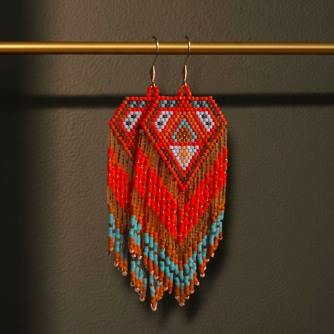 Handgewebte lange Perlenohrringe Rosso di Mare, aufgehängt an einer goldenen Präsentationsstange vor einem dunklen Hintergrund. Das Design zeigt ein intensives Korallenrot kombiniert mit kühlem Türkisblau und Sandtönen in einem symmetrischen Rautenmuster.