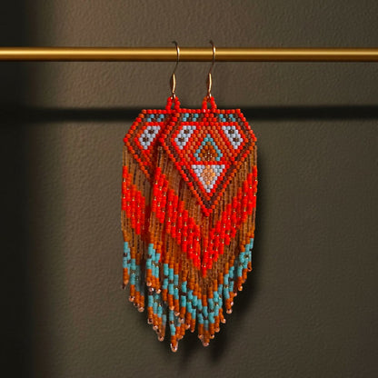 Handgewebte lange Perlenohrringe Rosso di Mare, aufgehängt an einer goldenen Präsentationsstange vor einem dunklen Hintergrund. Das Design zeigt ein intensives Korallenrot kombiniert mit kühlem Türkisblau und Sandtönen in einem symmetrischen Rautenmuster.