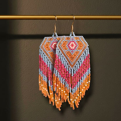 Handgewebte lange Perlenohrringe Sabbia Corallo, aufgehängt an einer filigranen goldenen Präsentationsstange vor einem dunklen, stimmungsvollen Hintergrund. Zu sehen ist das maritime Design in Koralle, Beerenrot und Hellblau mit fließenden Fransen.