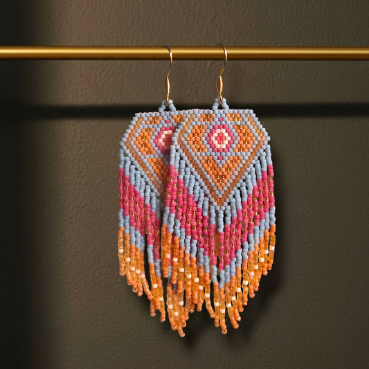 Handgewebte lange Perlenohrringe Sabbia Corallo, aufgehängt an einer filigranen goldenen Präsentationsstange vor einem dunklen, stimmungsvollen Hintergrund. Zu sehen ist das maritime Design in Koralle, Beerenrot und Hellblau mit fließenden Fransen.
