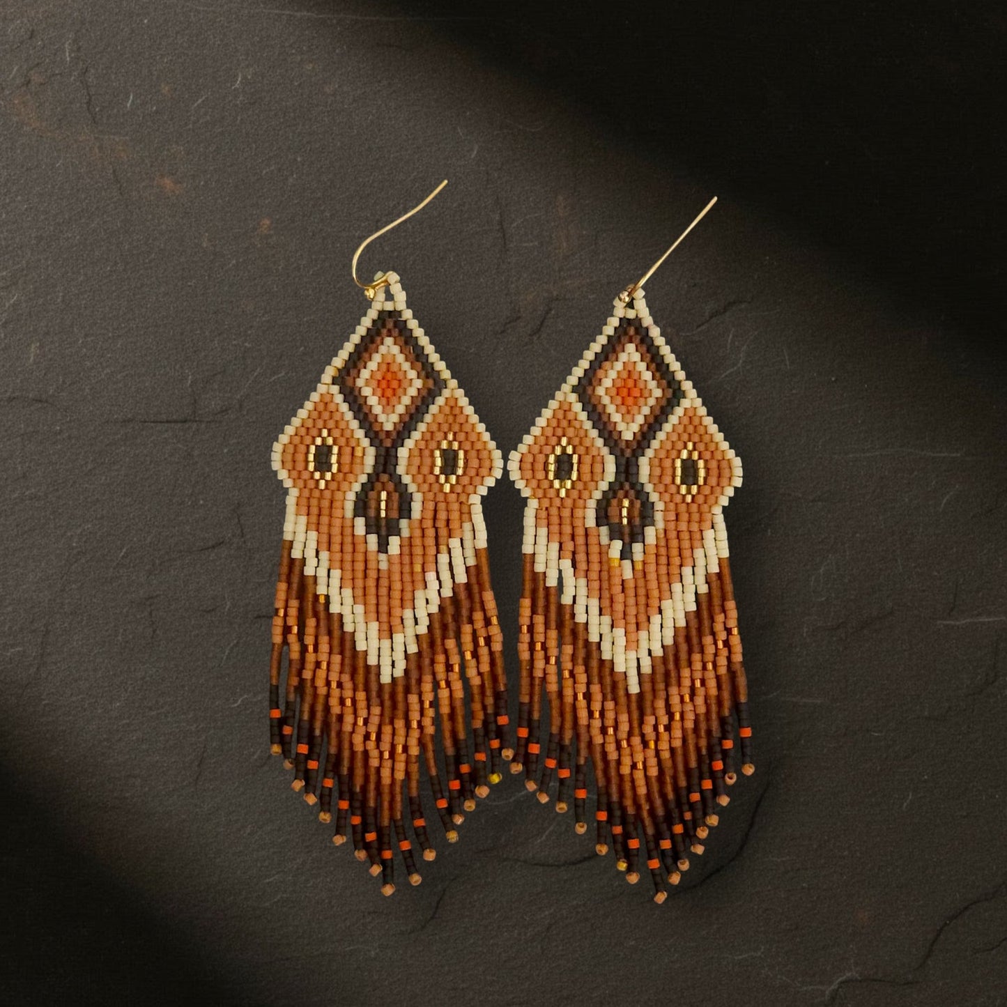 Handgefertigte Statement-Ohrringe mit geometrischem Rautenmuster aus japanischen Miyuki Delica Perlen in Orange, Terracotta, Schwarz und Beige.
Die langen Perlenfransen hängen an goldfarbenen Ohrhaken.