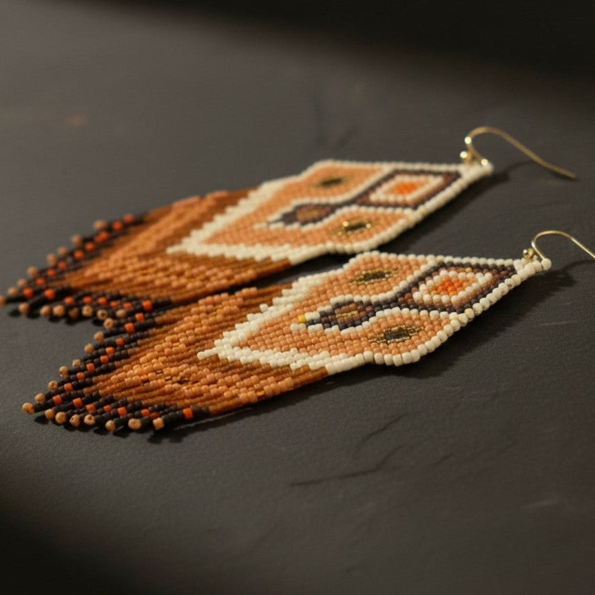 Eine detaillierte Makroaufnahme der "Miraggio Sabbioso" Ohrringe auf einer dunklen Schieferplatte. Die präzise gewebten Miyuki-Perlen zeigen ein kunstvolles Webmuster mit harmonischen Übergängen zwischen Creme, Goldbraun und tiefem Terrakotta.