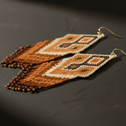 Eine detaillierte Makroaufnahme der "Miraggio Sabbioso" Ohrringe auf einer dunklen Schieferplatte. Die präzise gewebten Miyuki-Perlen zeigen ein kunstvolles Webmuster mit harmonischen Übergängen zwischen Creme, Goldbraun und tiefem Terrakotta.