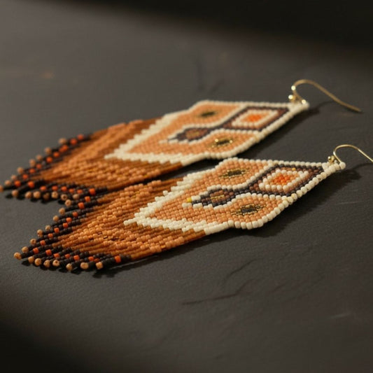 Eine detaillierte Makroaufnahme der "Miraggio Sabbioso" Ohrringe auf einer dunklen Schieferplatte. Die präzise gewebten Miyuki-Perlen zeigen ein kunstvolles Webmuster mit harmonischen Übergängen zwischen Creme, Goldbraun und tiefem Terrakotta.