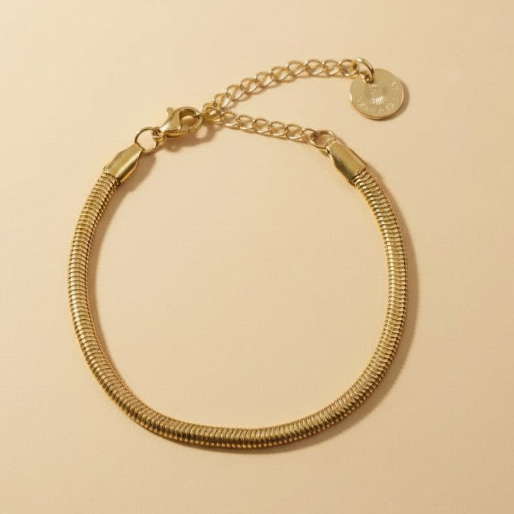 Goldfarbenes Serena Snake Armband mit flacher Edelstahlstruktur auf warmem beigem Hintergrund.