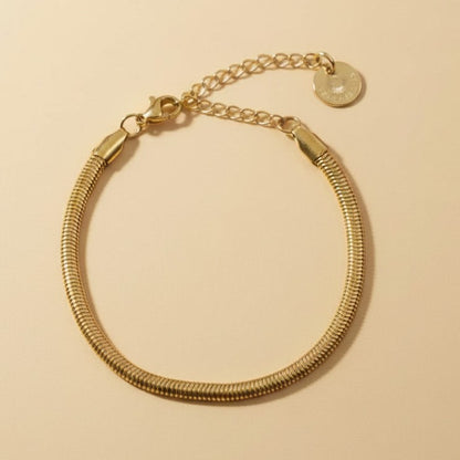Goldfarbenes Serena Snake Armband mit flacher Edelstahlstruktur auf warmem beigem Hintergrund.