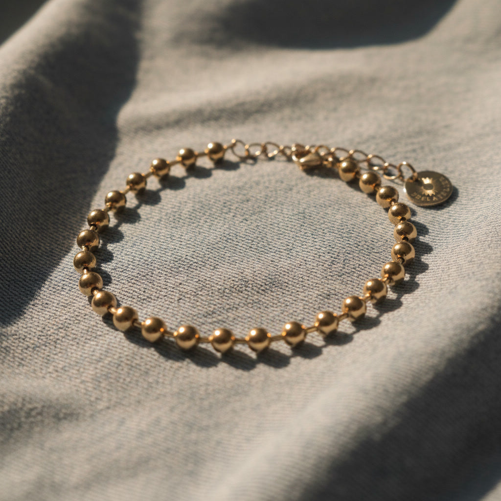 Vergoldetes Sfera Kugelarmband aus Edelstahl 304L auf hellblauem Denimstoff im Sonnenlicht.