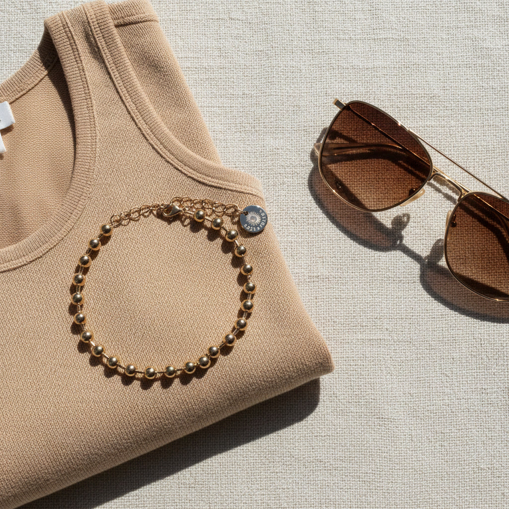 Sfera Kugelarmband aus vergoldetem Edelstahl auf beigem Oberteil neben einer Sonnenbrille im warmen Sommerlicht.