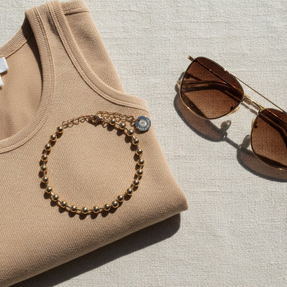 Sfera Kugelarmband aus vergoldetem Edelstahl auf beigem Oberteil neben einer Sonnenbrille im warmen Sommerlicht.