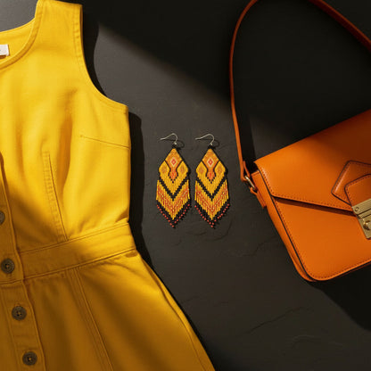 Zentral auf einer dunklen Schieferplatte liegen die Perlenohrringe in Braun, Orange und Goldbraun. Links davon liegt eine edle orangefarbene Lederhandtasche mit goldenem Verschluss. Auf der rechen Seite liegt ein tolles, knallgelbes Retrojeanskleid. Ein Lichtstrahl setzt die Szene diagonal in Szene.